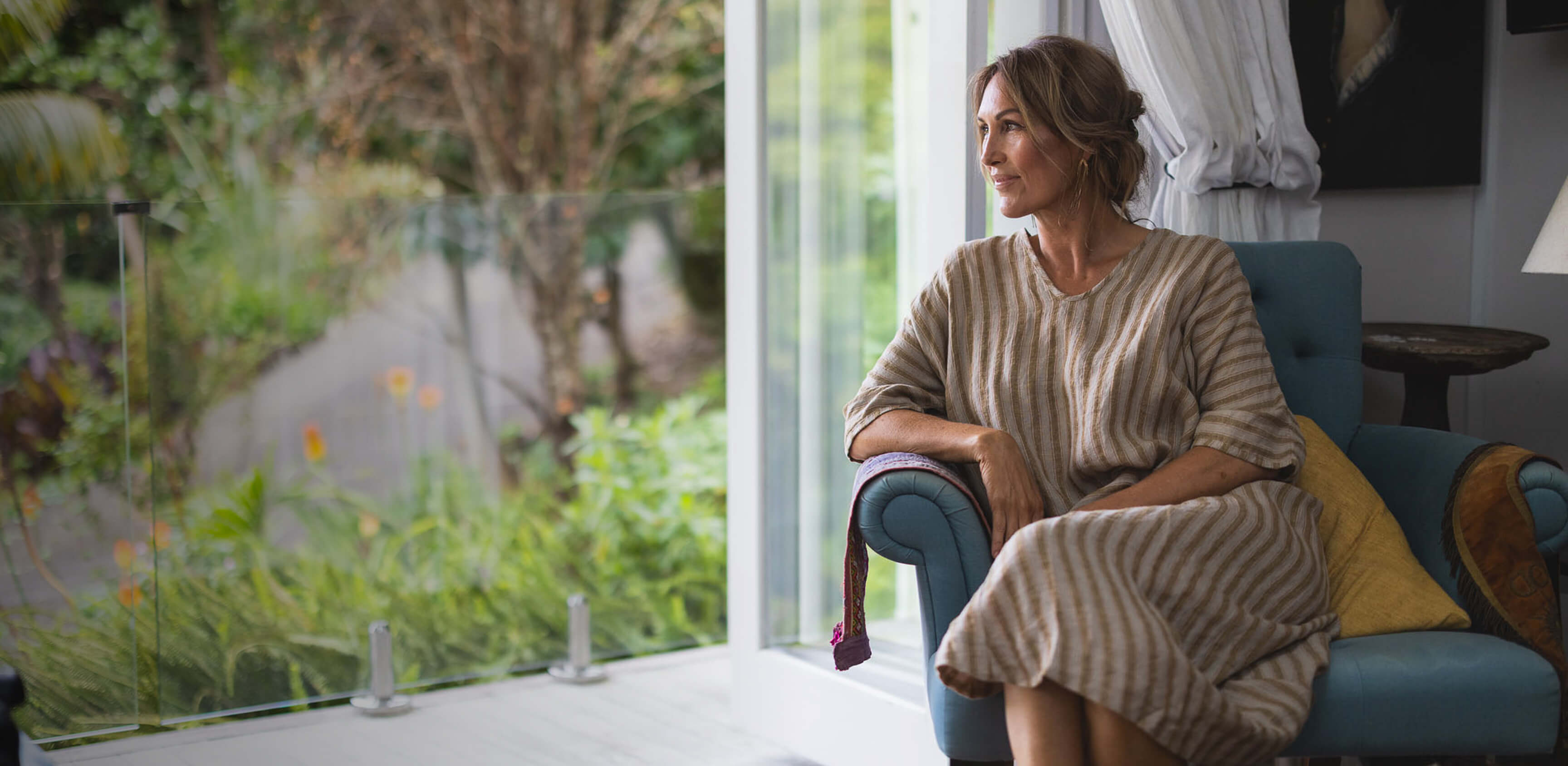 Woman sitting on a blue chair in a room with a garden view wearing The Tailor and his Lover dress