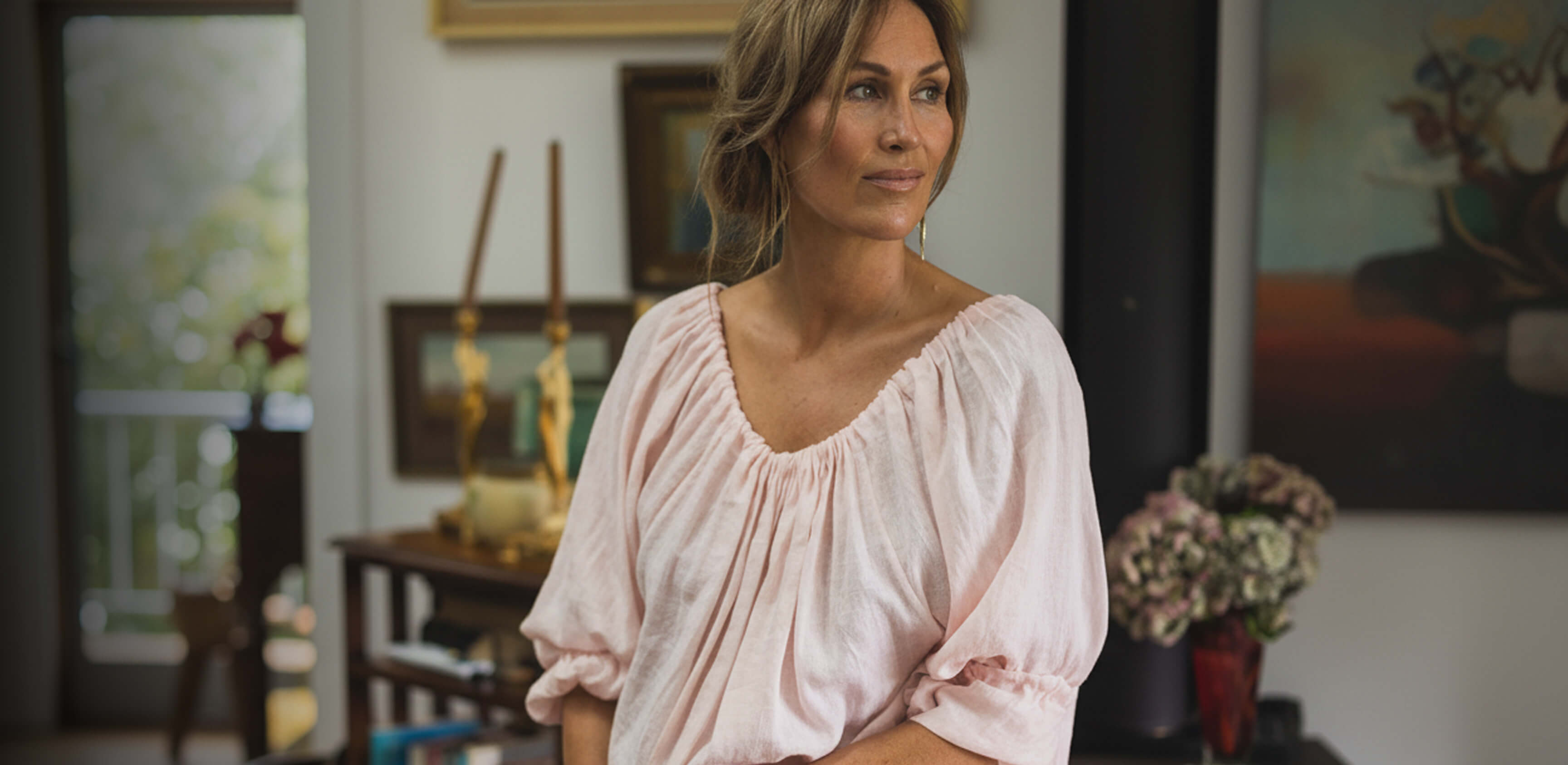 Woman in a pink The Tailor and his Lover blouse standing in a room with decor elements.